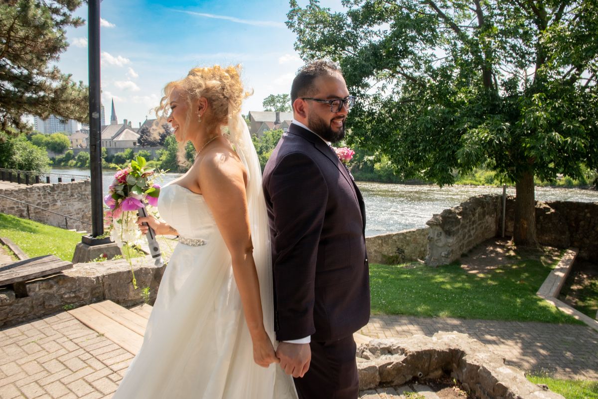 A smiling bride and groom hold hands along a stone path beside a river, with greenery and cityscape in the background.