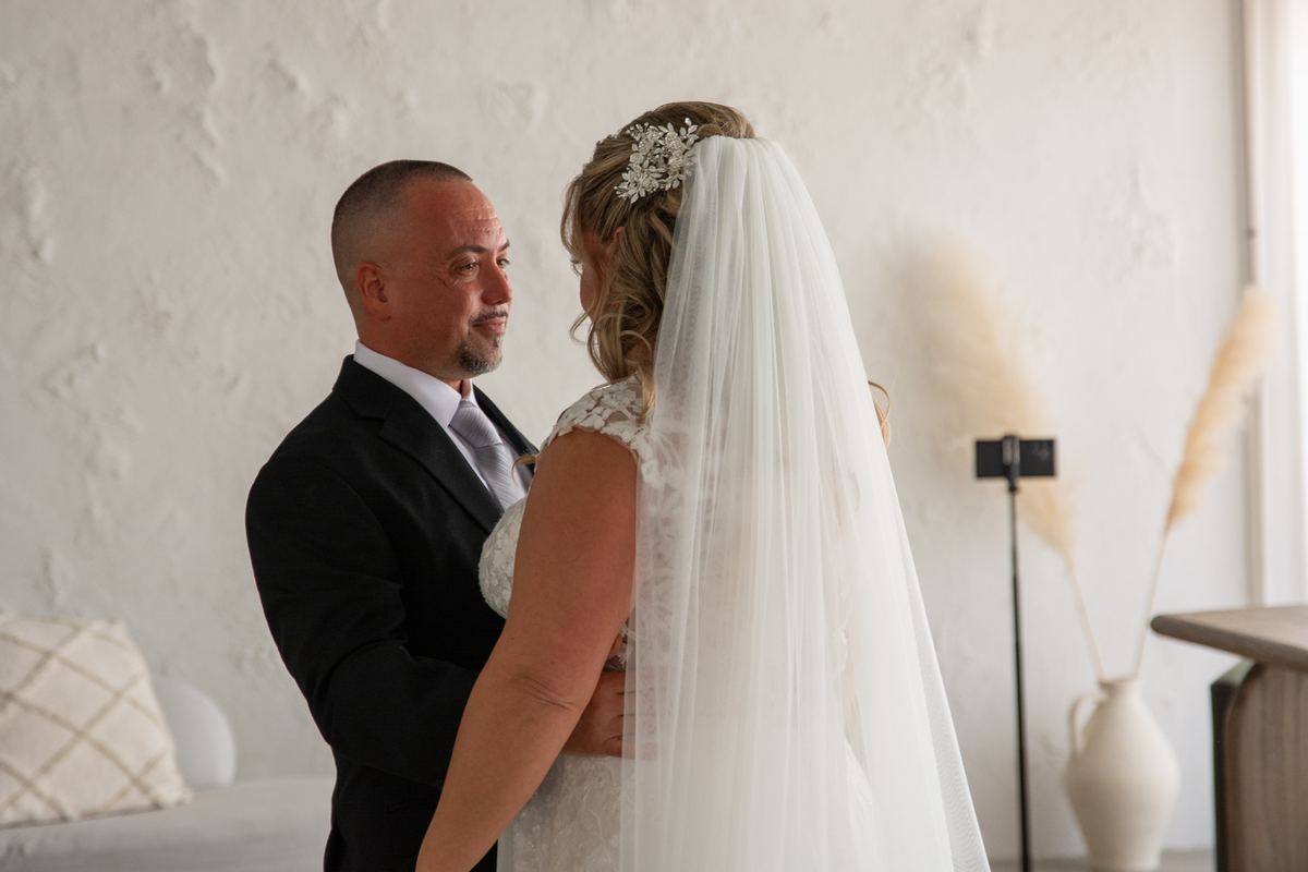 A groom embraces his bride from behind in soft light, their eyes meeting intimately against a white wall.
