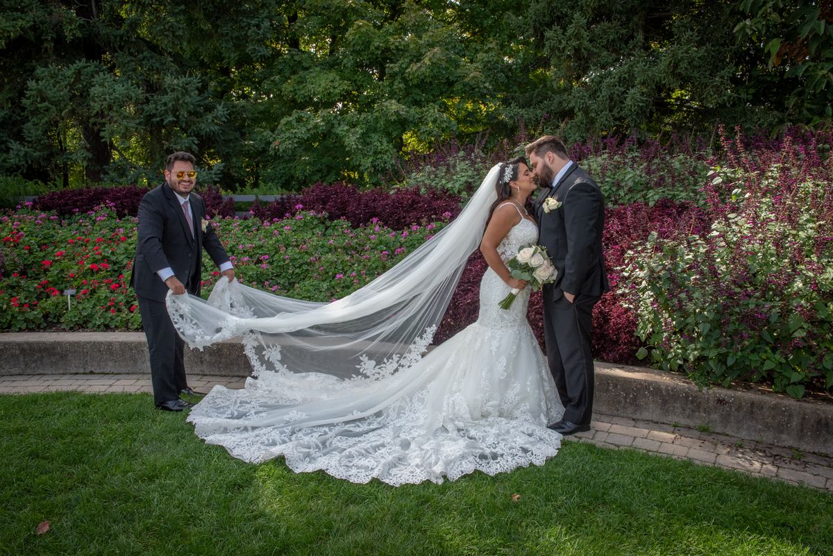 A bride and groom kiss as he holds her lace veil taut, framed by sunlight through trees and colorful flowers.