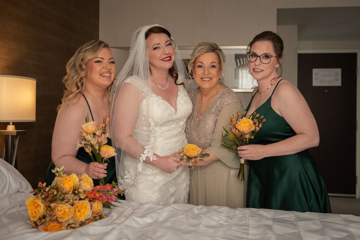 A bride and three women in formal attire stand smiling with floral bouquets near a bed covered in flowers.