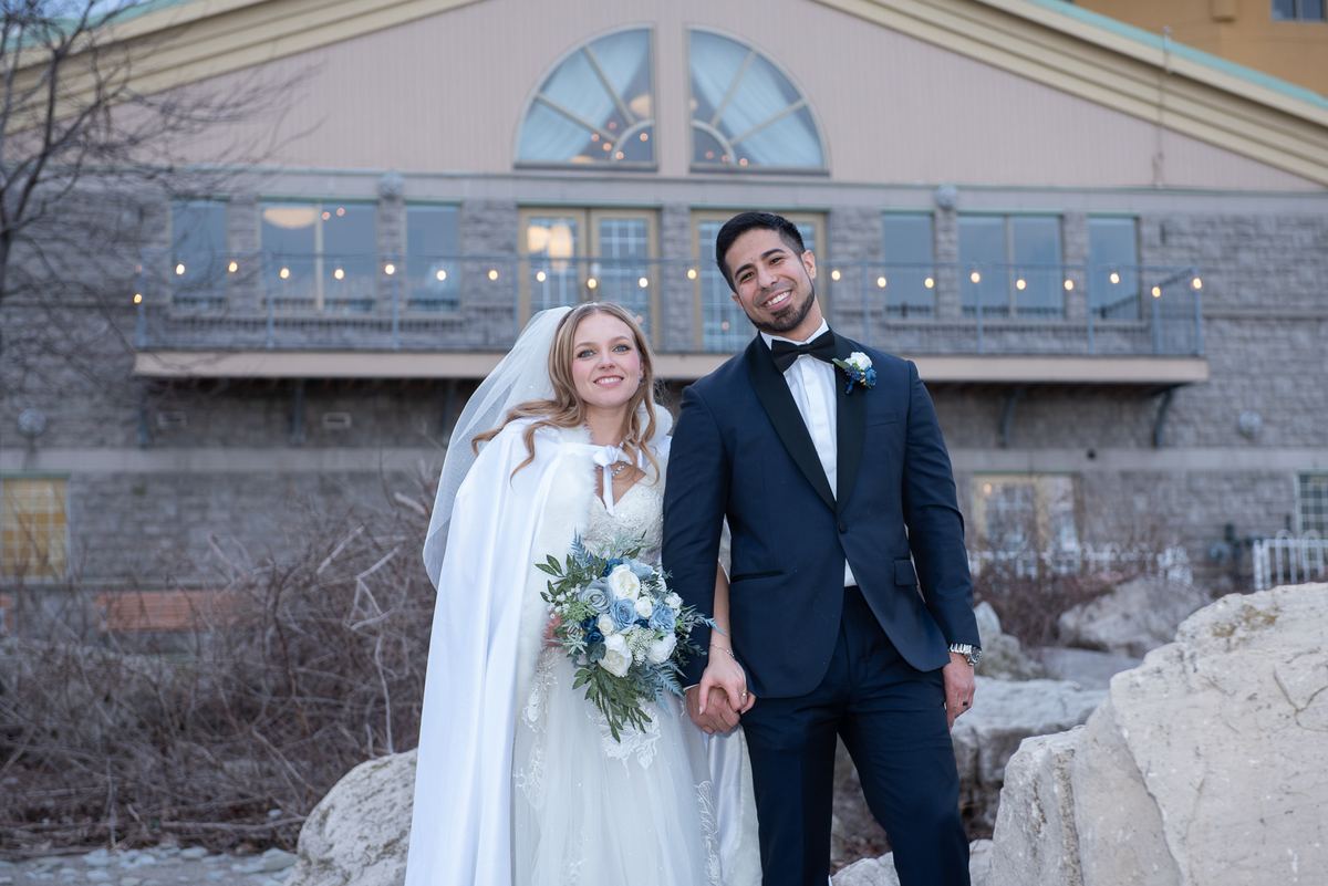 A smiling bride in a white gown and veil holds hands with her tuxedo-clad groom in front of an illuminated stone building.