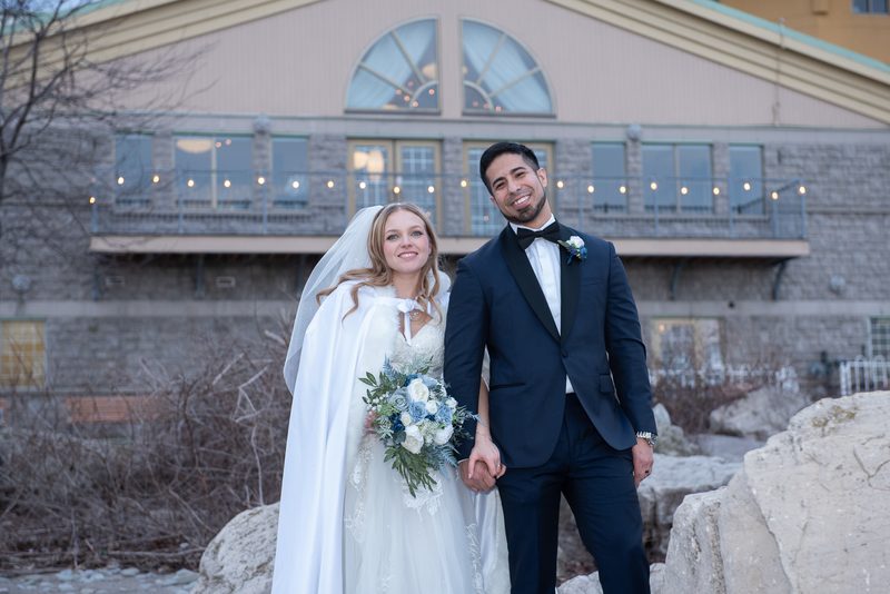 A smiling bride in a white gown and veil holds hands with her tuxedo-clad groom in front of an illuminated stone building.