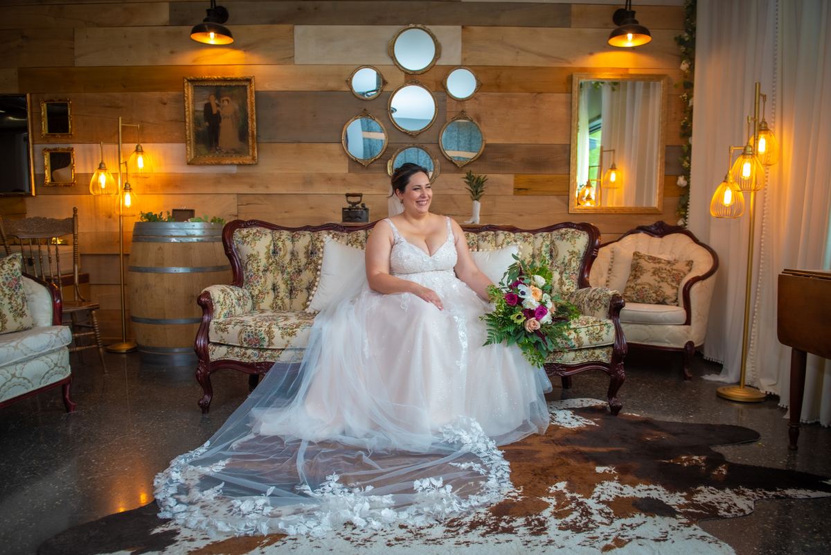 A smiling bride sits on a floral sofa in a rustic lounge with wooden walls and vintage lamps, holding a bouquet.
