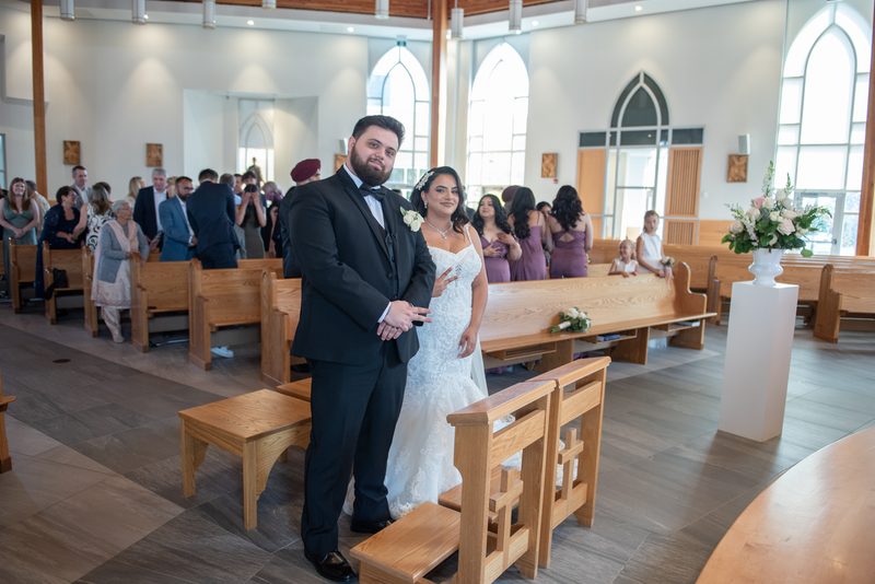 A bride and groom smile at the altar as guests sit in wooden pews, with natural light streaming through arched windows.