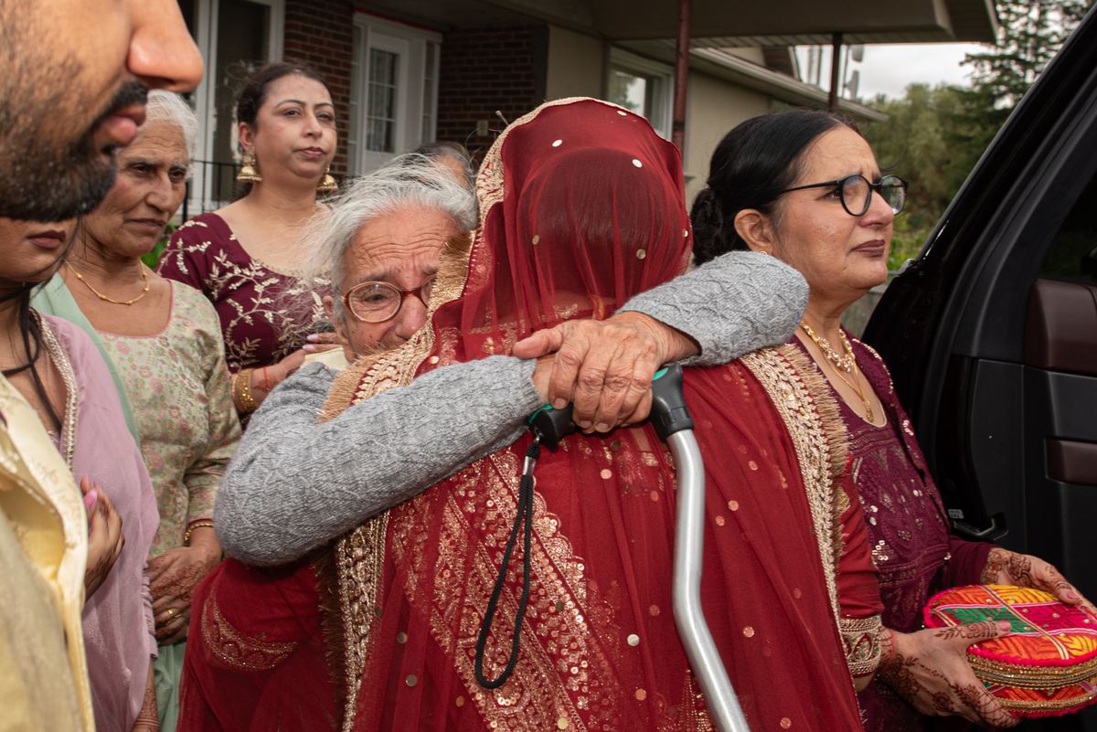 An elderly woman in red and gold is embraced by a younger woman with a walker as other women in traditional attire observe.