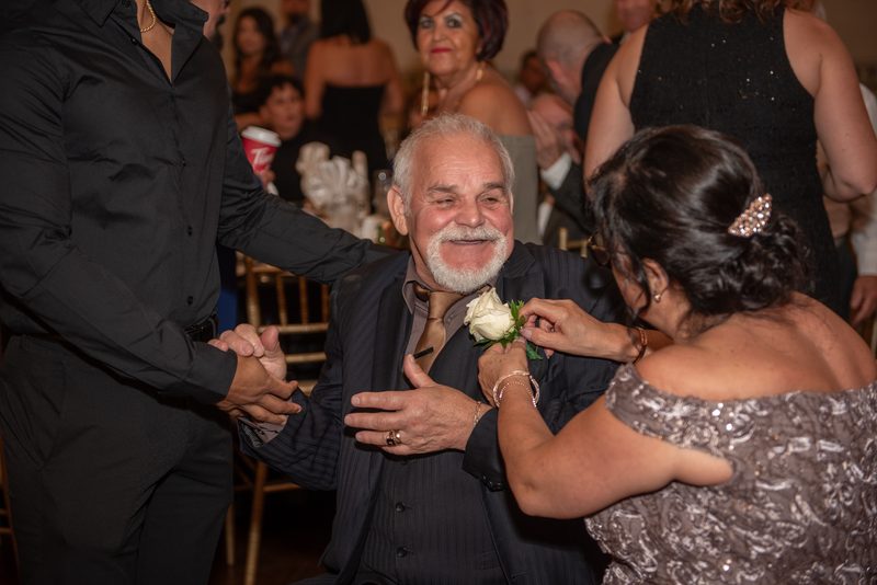 An older man in a pinstripe suit smiles as a woman pins a white rose boutonnière on him, another person holds his hands.