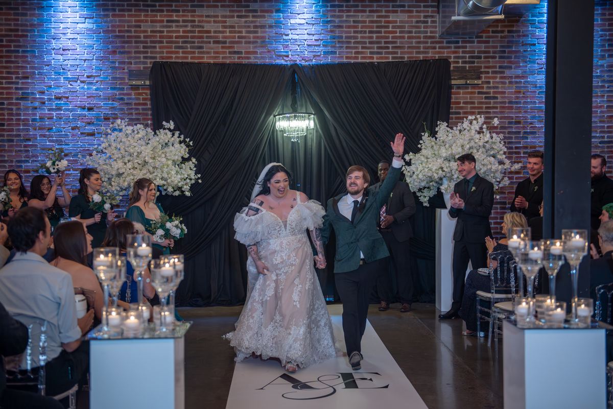 Bride and groom run down the aisle, arms raised, as guests applaud, with blue uplighting on brick wall.