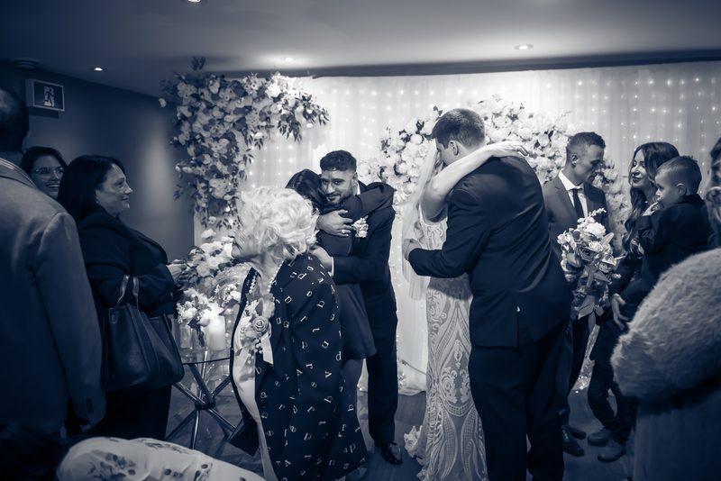 Bride and groom embrace guests, including an older woman, under string lights behind a floral arch at their reception.