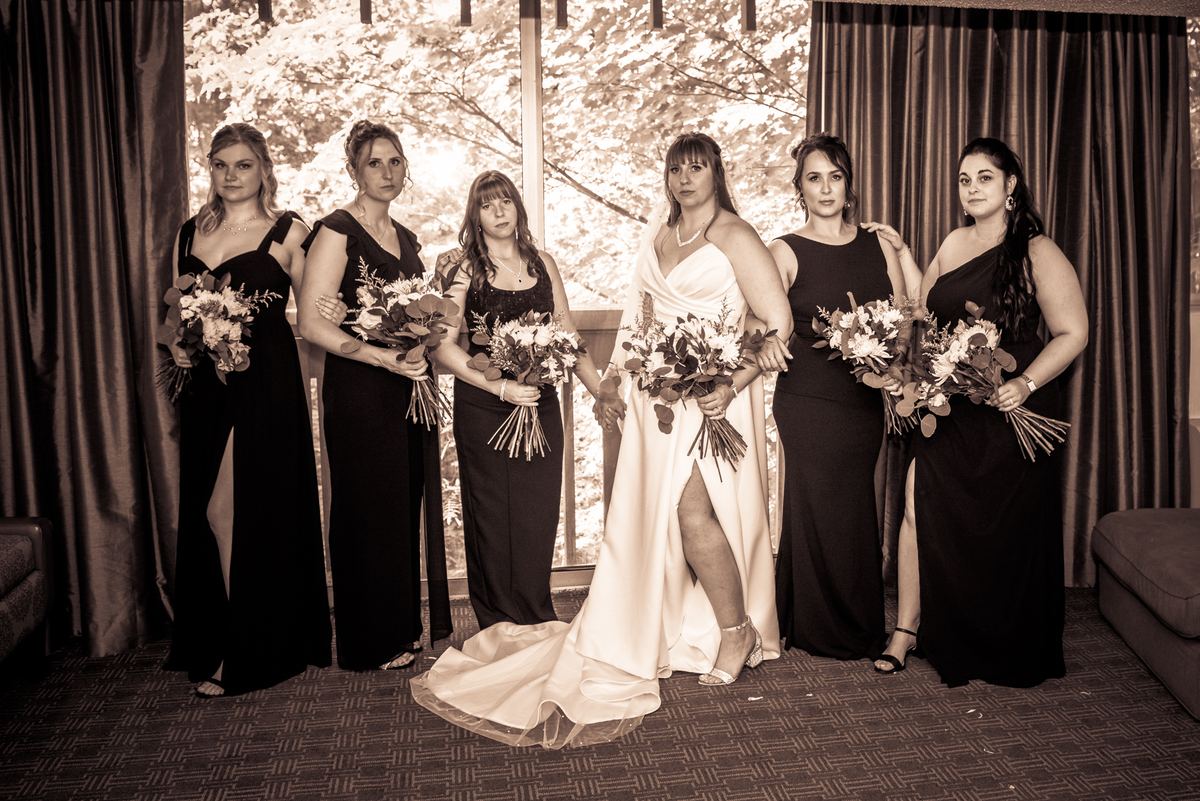 A bride in a white gown stands with her bridesmaids holding bouquets near a window letting in soft natural light.