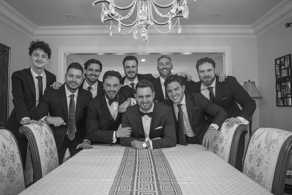 Groom and his attendants gather around a dining table for a warm, celebratory group portrait under an ornate chandelier.