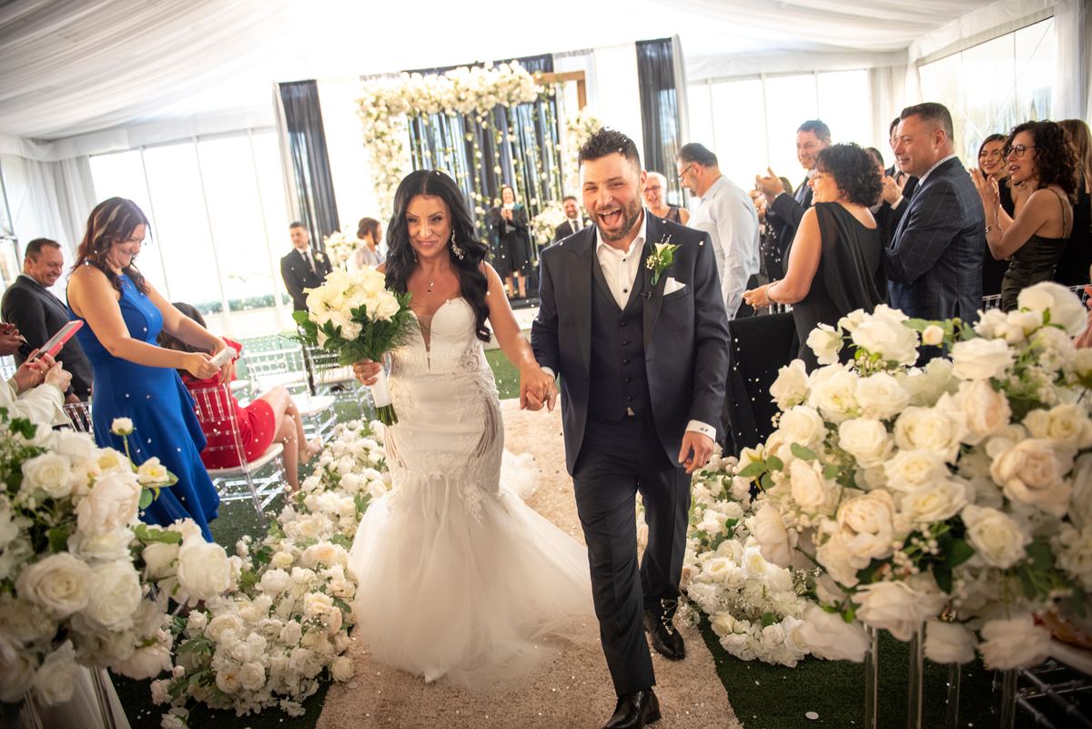 A smiling bride and groom walk down an aisle lined with white roses under a bright tent, surrounded by guests.