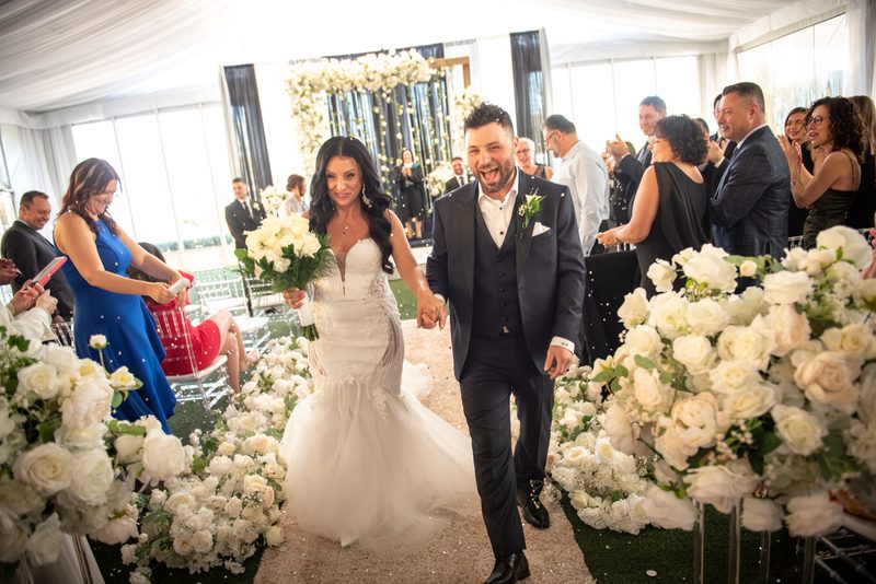A smiling bride and groom walk down an aisle lined with white roses under a bright tent, surrounded by guests.
