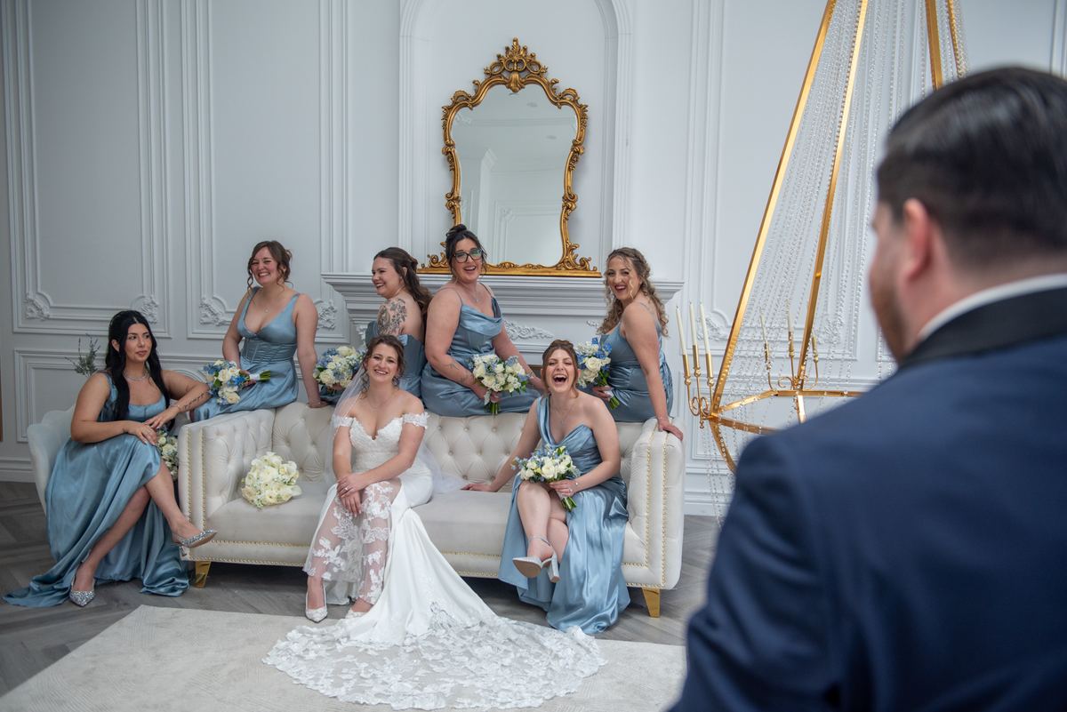 A bride and her bridesmaids pose with bouquets on an ornate sofa, smiling at the groom in the background.