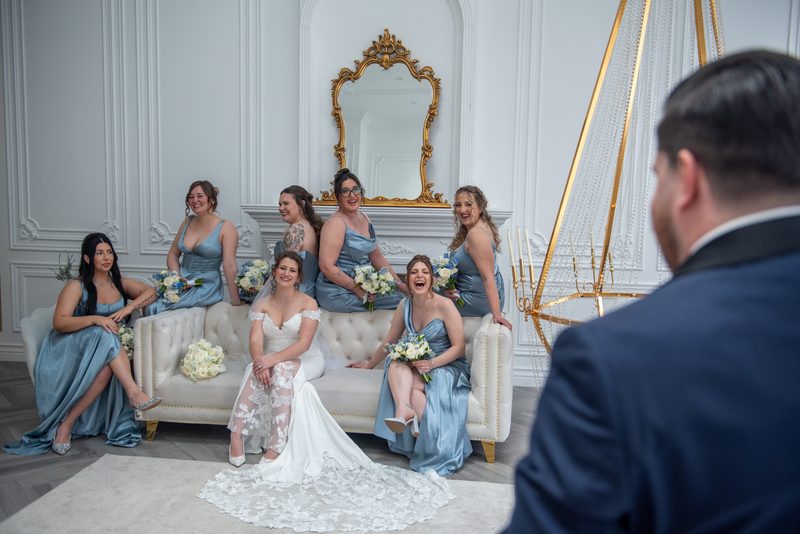 A bride and her bridesmaids pose with bouquets on an ornate sofa, smiling at the groom in the background.