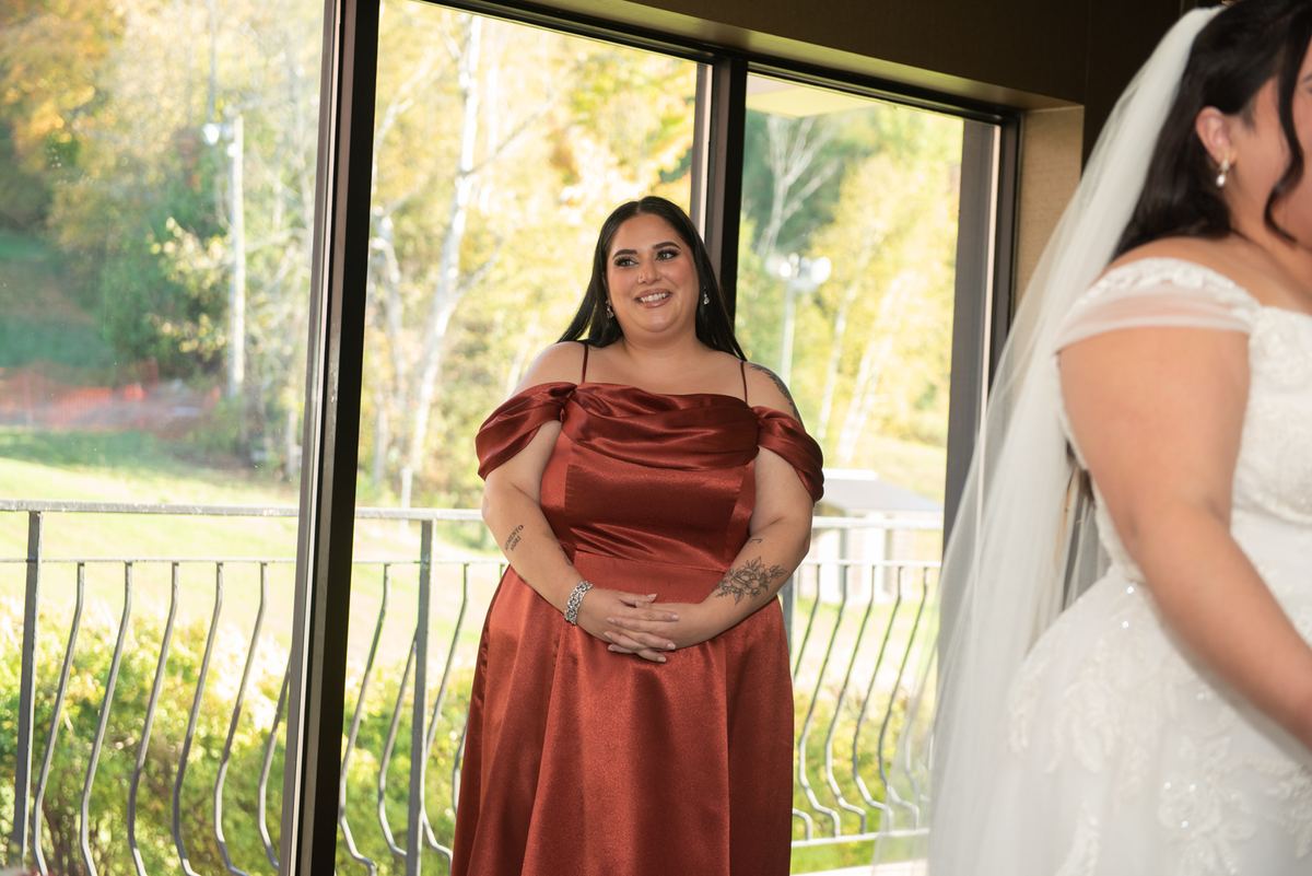A bridesmaid in a rust-colored gown smiles warmly at the bride in white, her hands clasped over her belly.