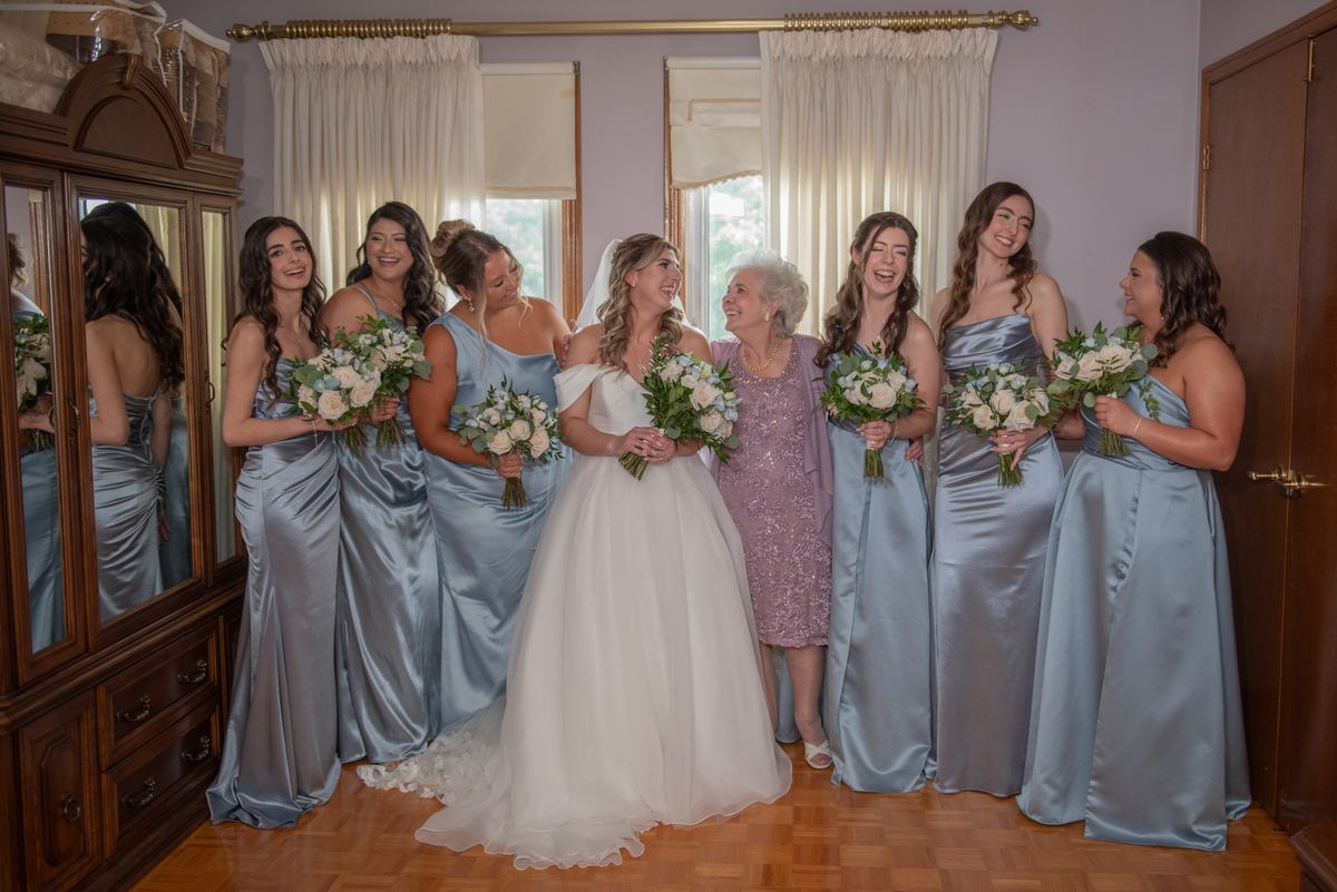 Bride in white gown surrounded by laughing bridesmaids in light blue gowns, holding flowers, with wooden floor.