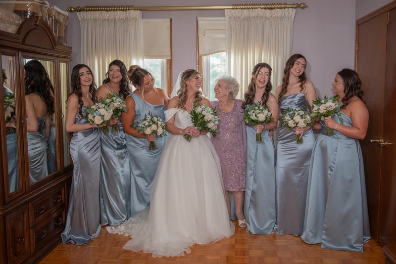 Bride in white gown surrounded by laughing bridesmaids in light blue gowns, holding flowers, with wooden floor.