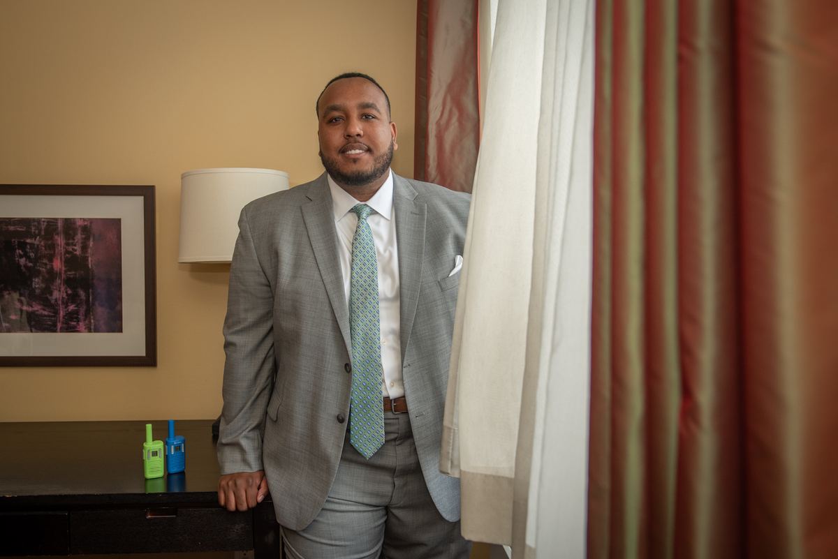 A man in a gray suit smiles next to a table with Hanukkah dreidels in a hotel room, suggesting celebration.