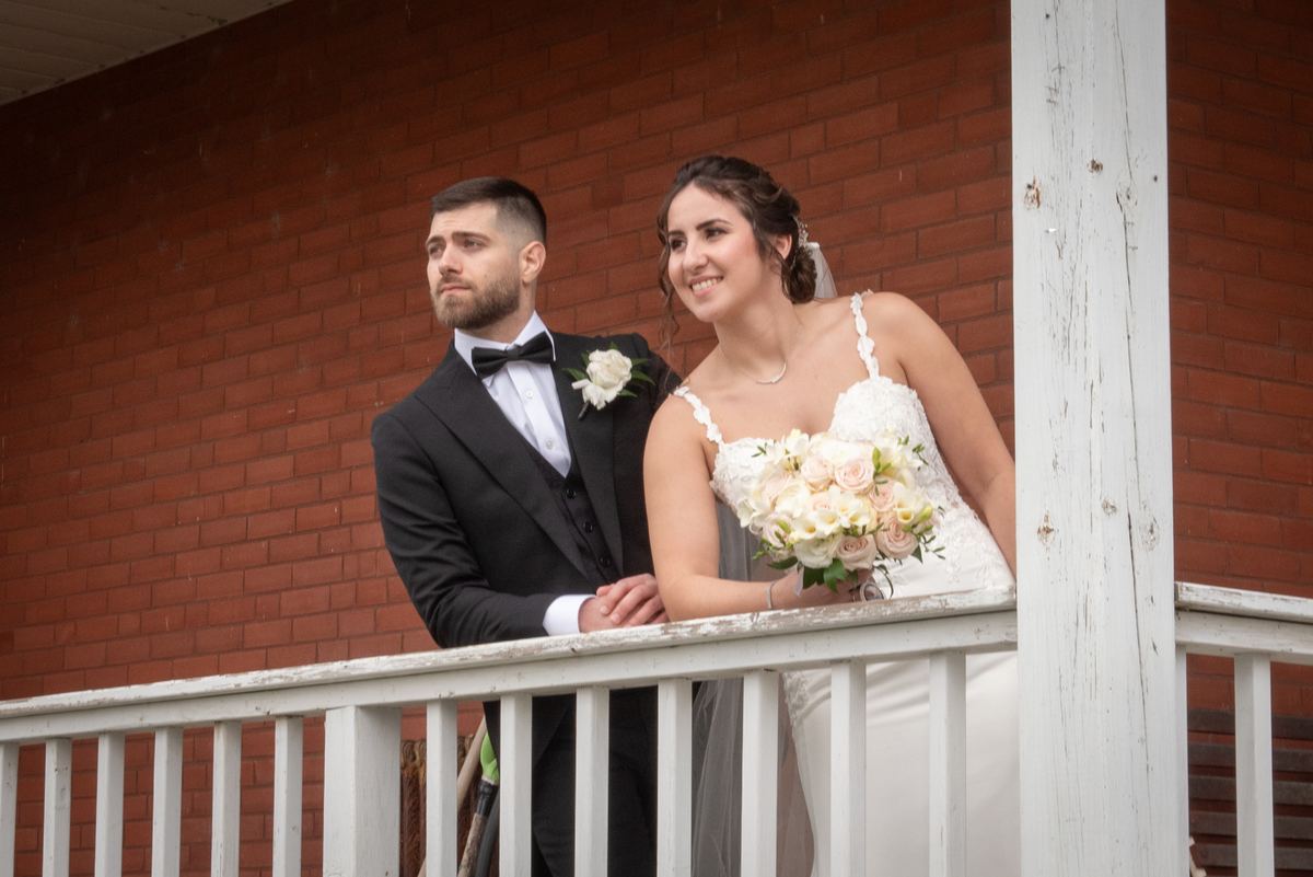 A newlywed couple in formal attire stands on a white porch railing, gazing out with joy against a red brick wall.