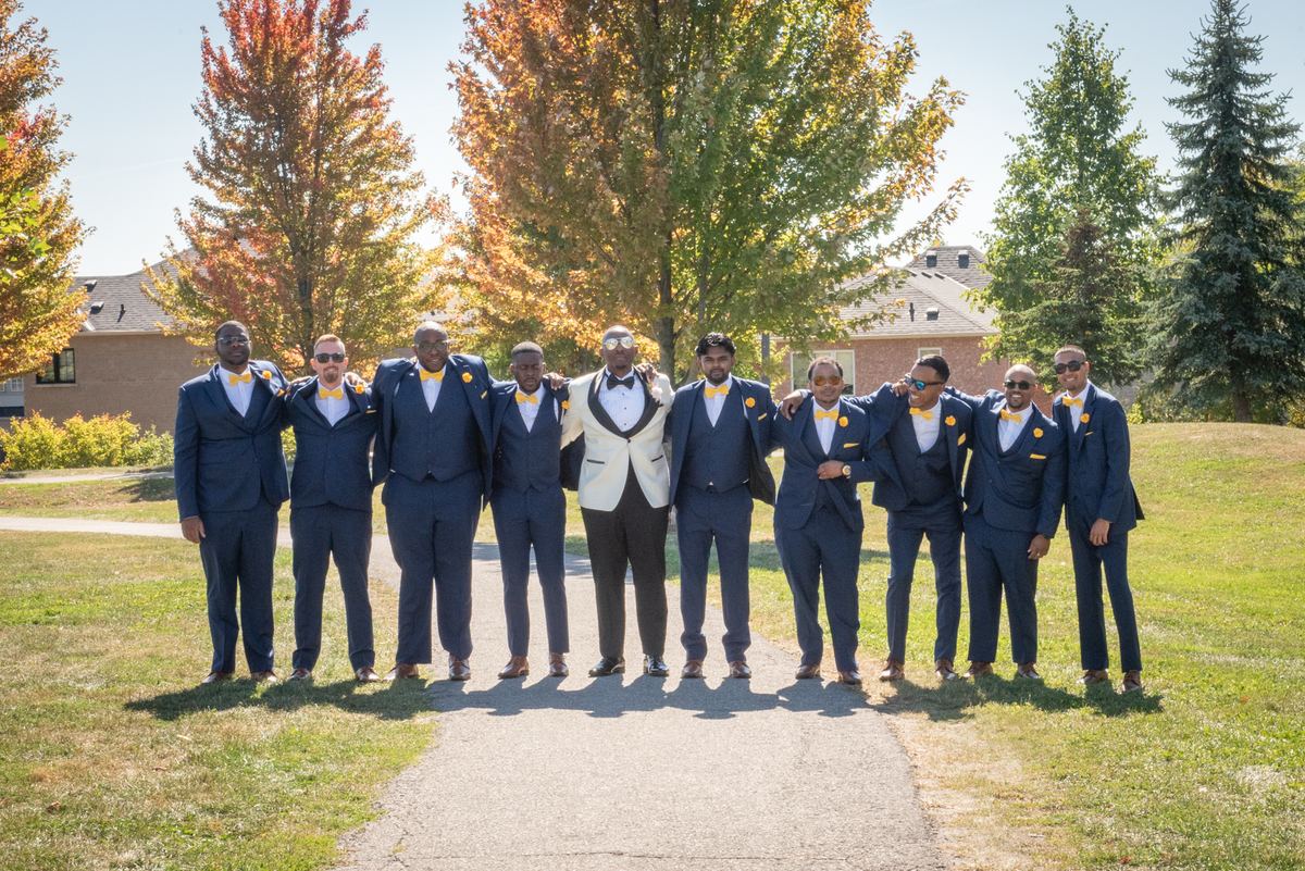 A groom in a white tuxedo and his navy-suited, yellow-bow-tied groomsmen pose on a path surrounded by fall foliage.