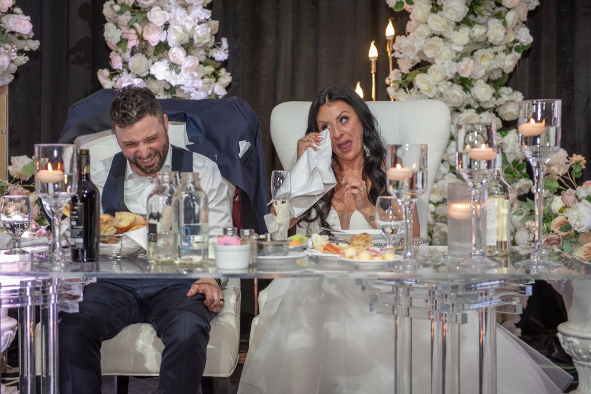 Bride wiping tears with napkin while groom laughs beside her at glass table set with candles and flowers.