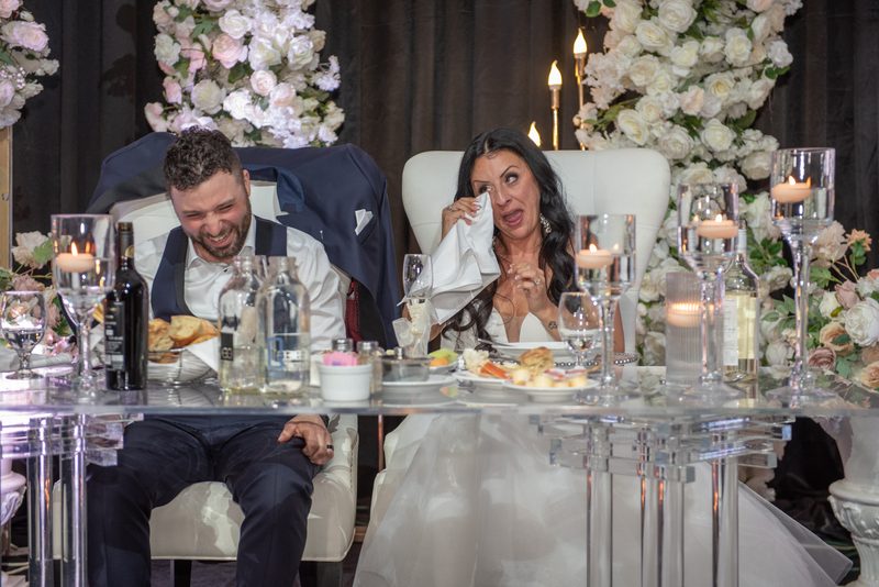 Bride wiping tears with napkin while groom laughs beside her at glass table set with candles and flowers.