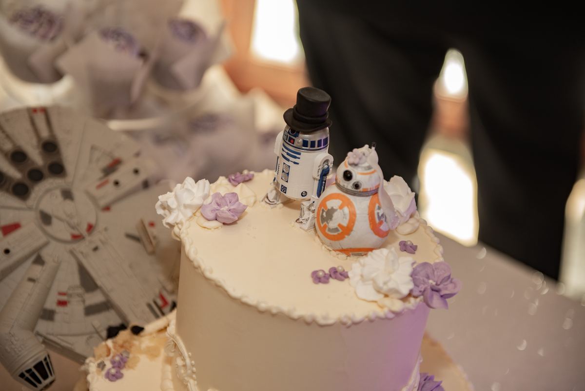 A wedding cake with R2-D2 and BB-8 as toppers in bride and groom outfits, near a Millennium Falcon model.