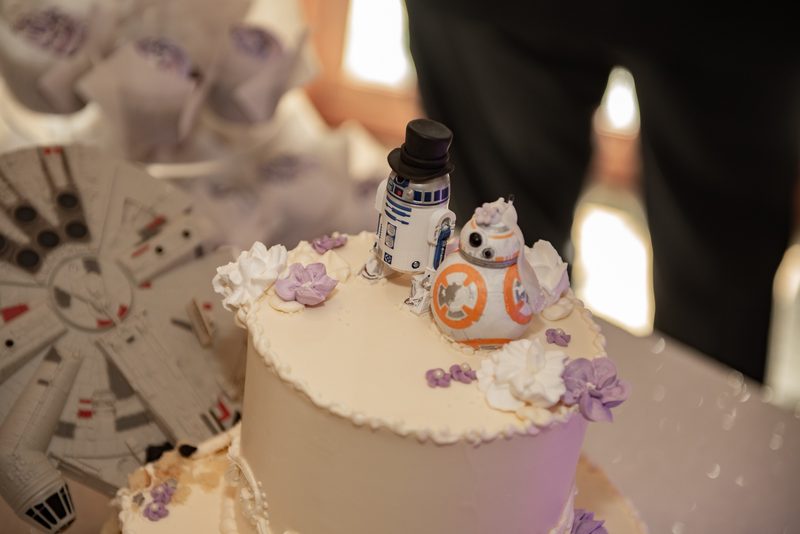 A wedding cake with R2-D2 and BB-8 as toppers in bride and groom outfits, near a Millennium Falcon model.