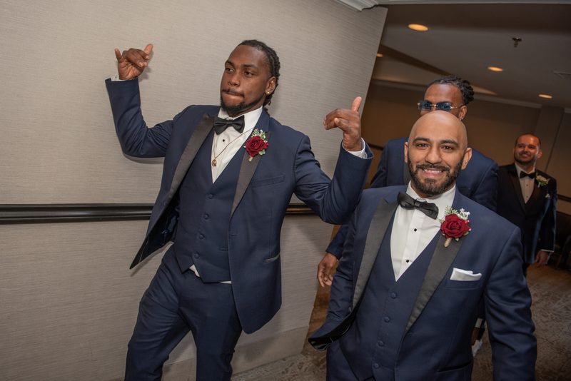 Groomsmen in navy tuxedos with red boutonnieres pose energetically at a reception, thumbs up and smiling.