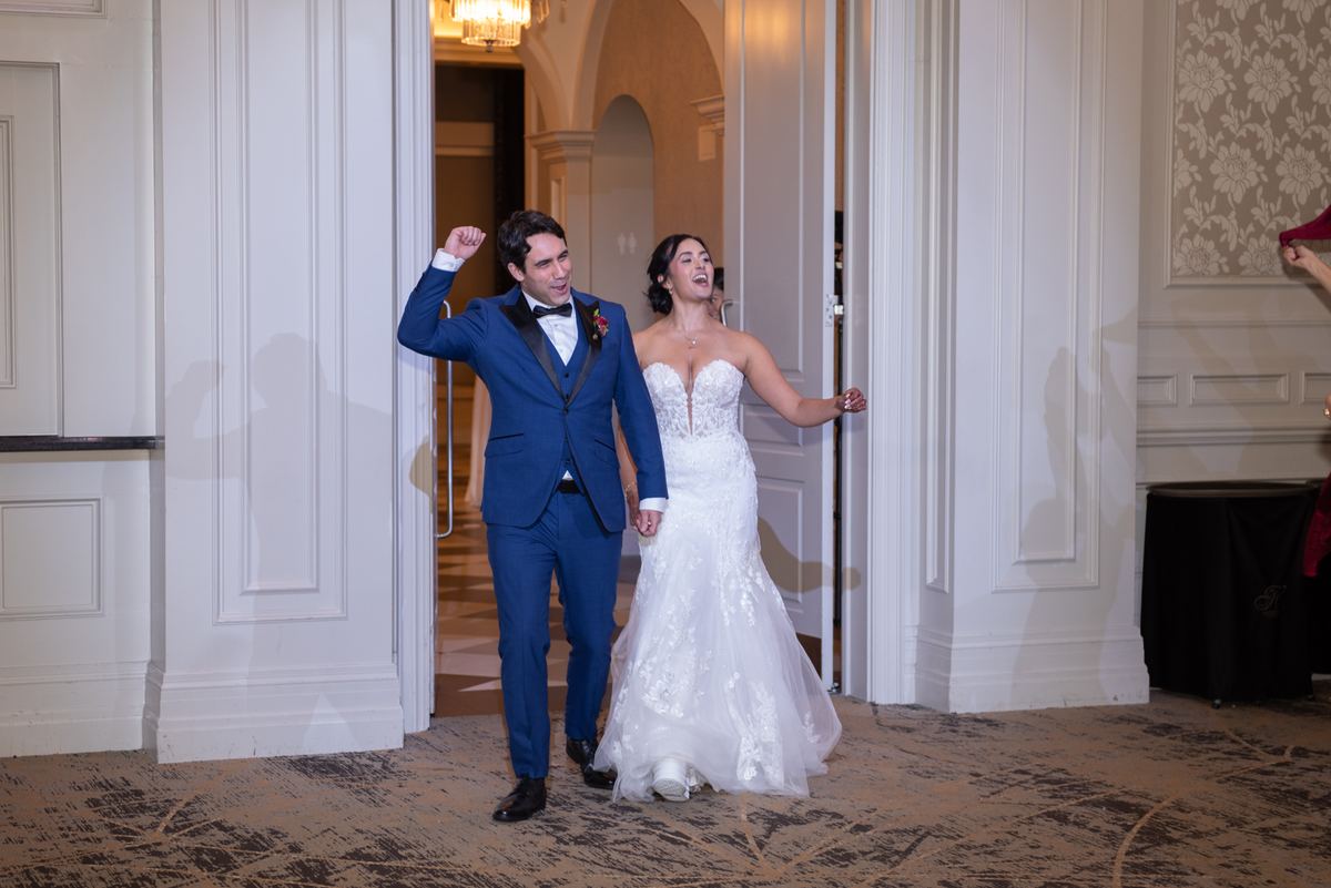 A bride and groom joyfully enter a grand hall through an ornate doorway, the warm light highlighting their exuberance.