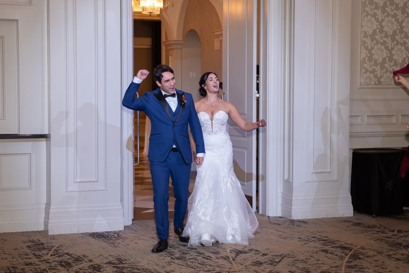 A bride and groom joyfully enter a grand hall through an ornate doorway, the warm light highlighting their exuberance.