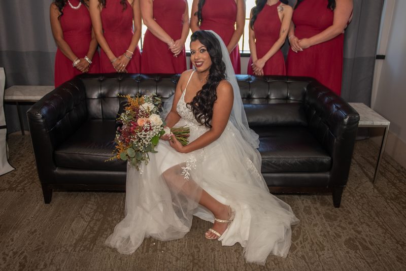 A bride sits on a leather couch with a bouquet, smiling at the camera as her bridesmaids stand behind her.