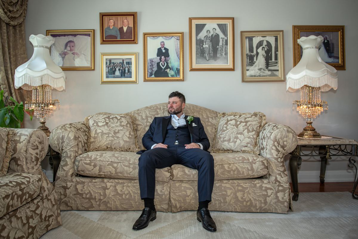 A groom in a navy tuxedo sits on a floral sofa, looking thoughtful amid family photos and crystal lamps.