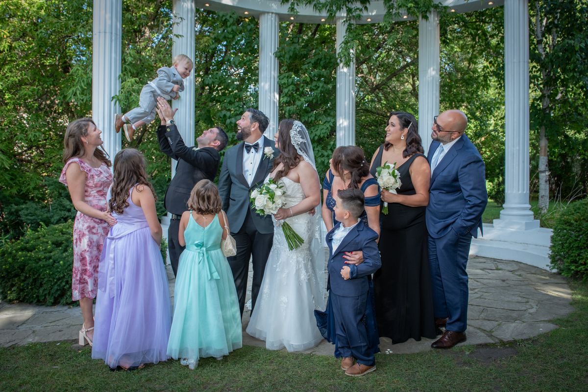 Groom lifts a child under a white gazebo, surrounded by smiling guests in formal attire, bathed in natural daylight.