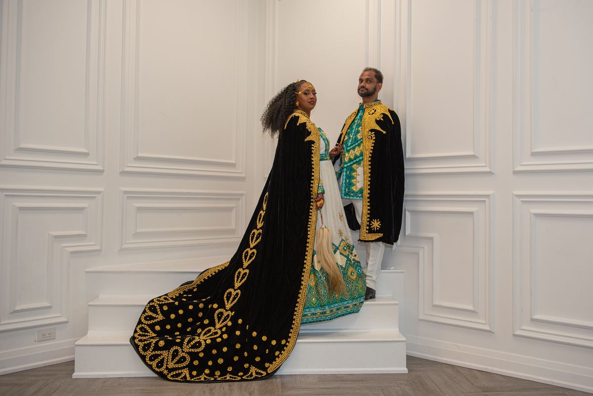 A couple in ornate black and gold ceremonial attire stands gracefully on white stairs, illuminated by soft light.