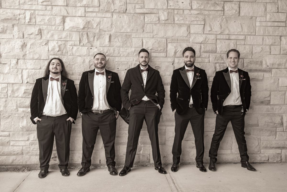 Five formally dressed men in tuxedos lean casually against a stone wall, exuding relaxed camaraderie.