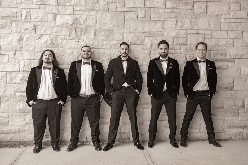 Five formally dressed men in tuxedos lean casually against a stone wall, exuding relaxed camaraderie.