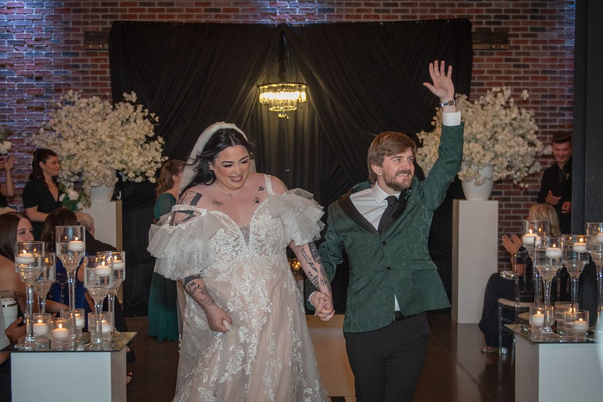 Bride in lace dress with tattoos and groom in green velvet jacket wave to guests at candlelit wedding.