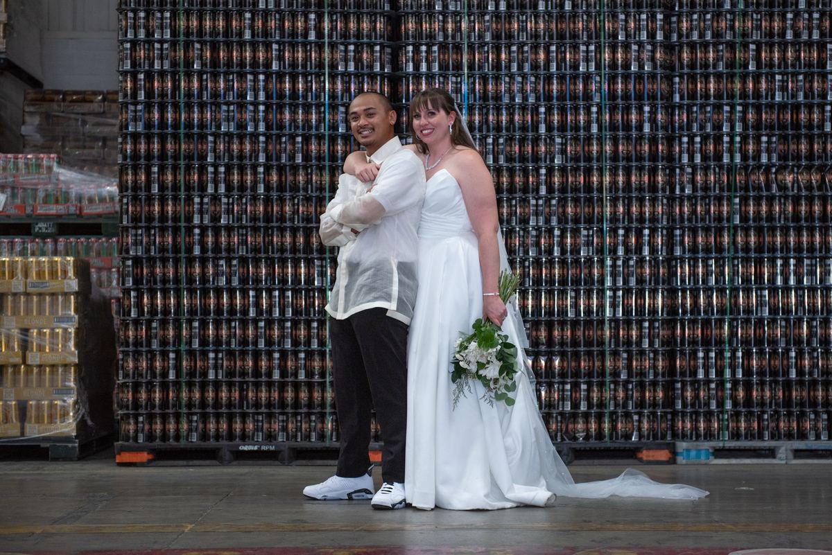 Samantha and Robbie stand joyfully in front of beer can pallets at their wedding, dressed in traditional attire.