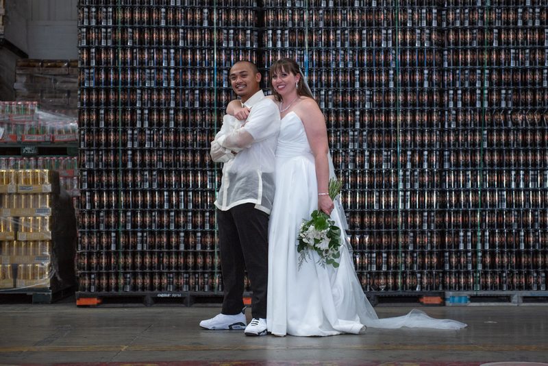 Samantha and Robbie stand joyfully in front of beer can pallets at their wedding, dressed in traditional attire.