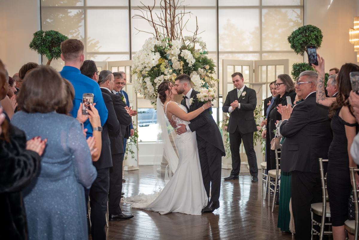 Bride and groom kiss at altar, surrounded by applauding guests and natural light streaming through windows.
