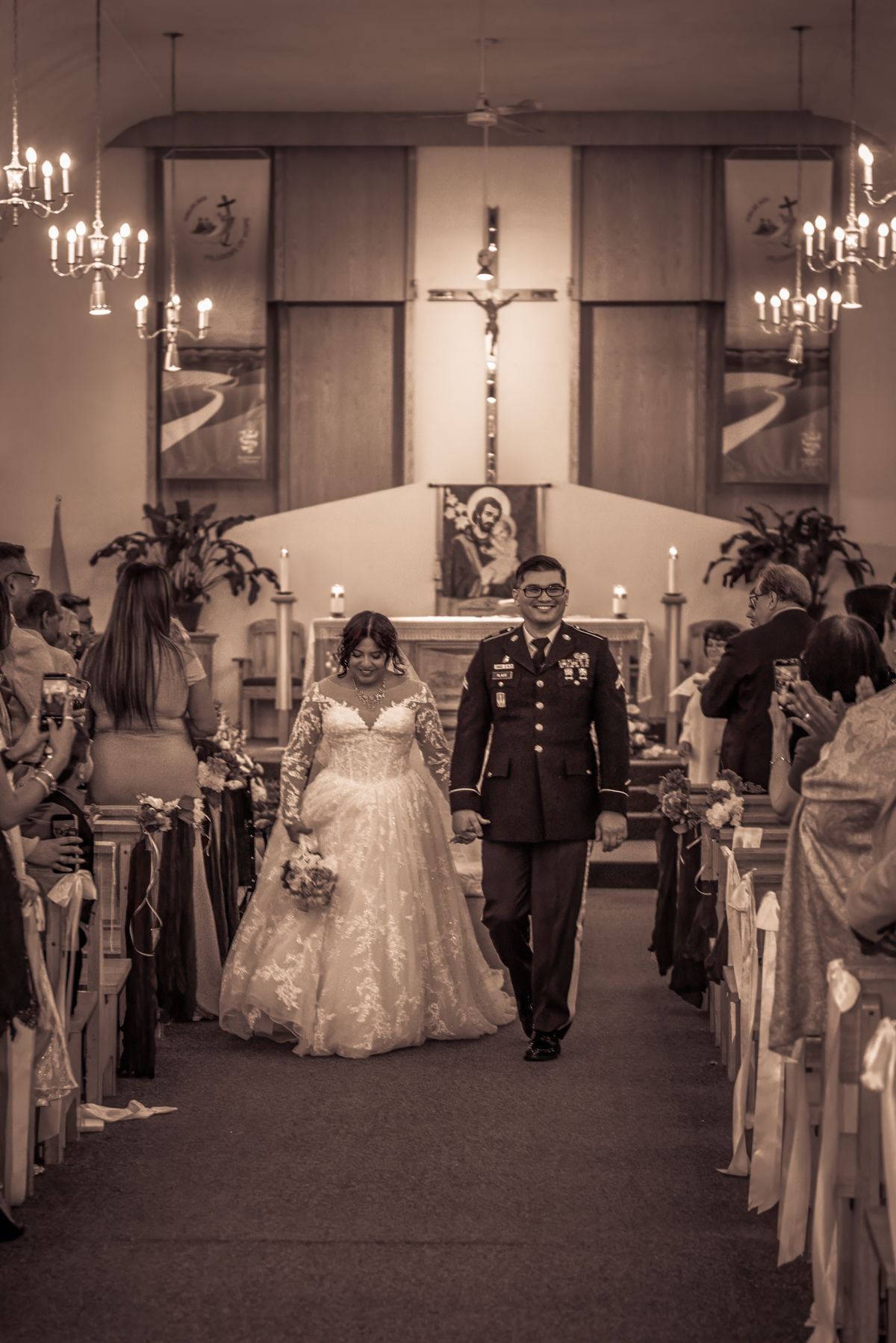 A bride in a lace gown and groom in military uniform walk down the church aisle, holding hands and smiling.