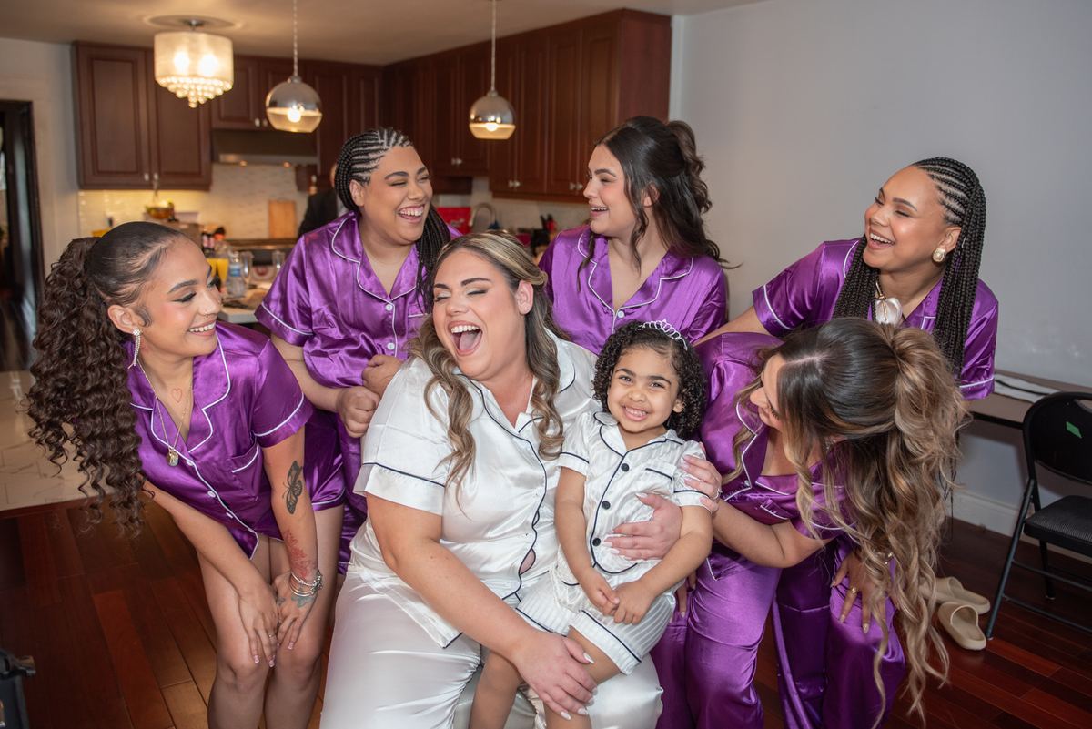 A bride in white satin pajamas laughs with her bridesmaids and a young girl on her lap in a warmly lit kitchen.