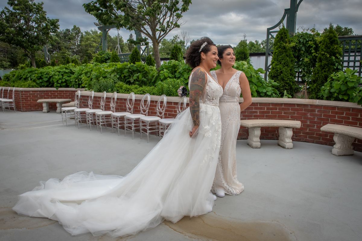 Two brides in white gowns stand on a patio, smiling at the camera near rows of empty chairs and greenery.