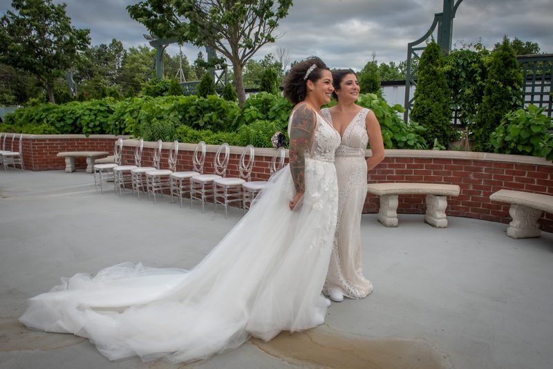 Two brides in white gowns stand on a patio, smiling at the camera near rows of empty chairs and greenery.