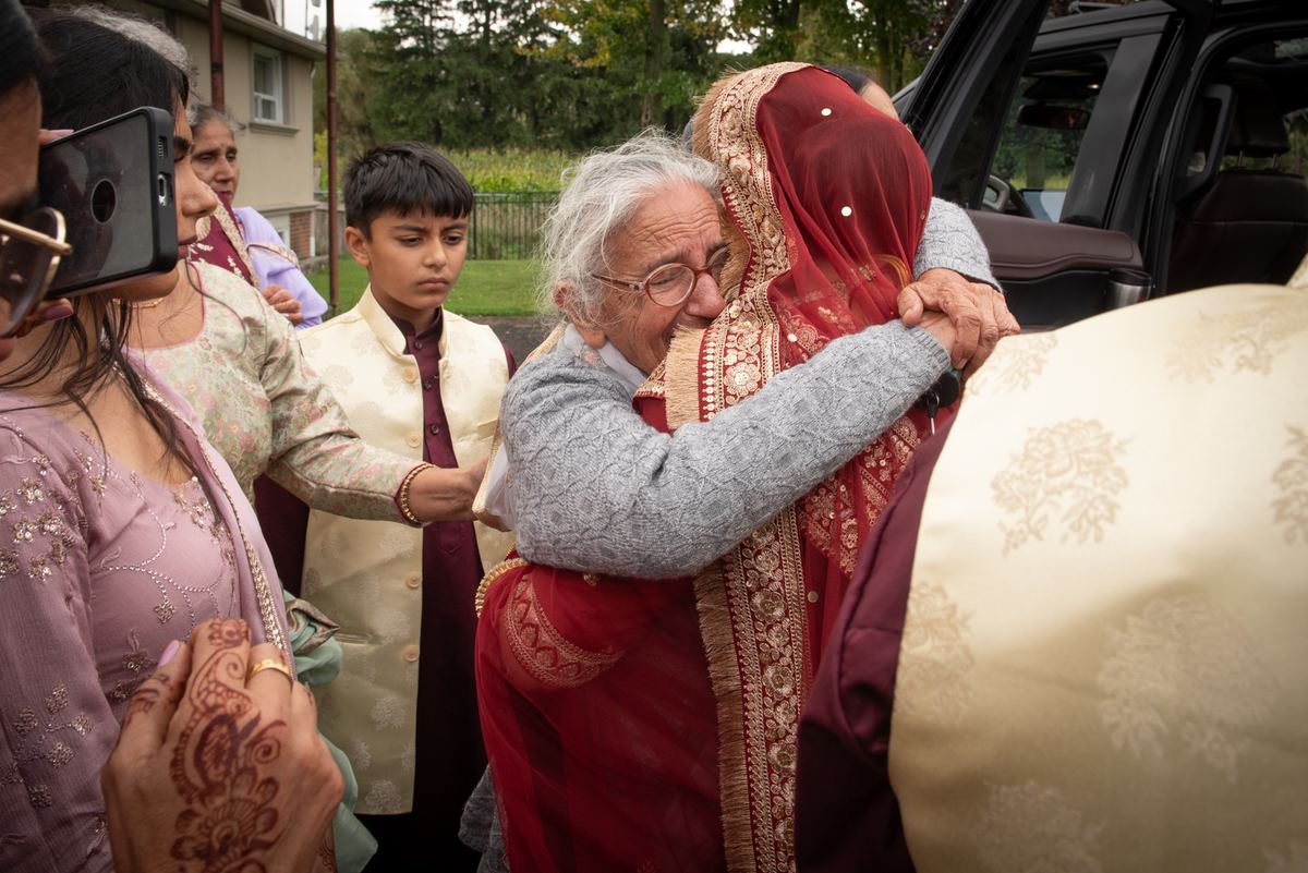 An elderly woman embraces a bride in a red and gold sari, surrounded by family members at an outdoor event.