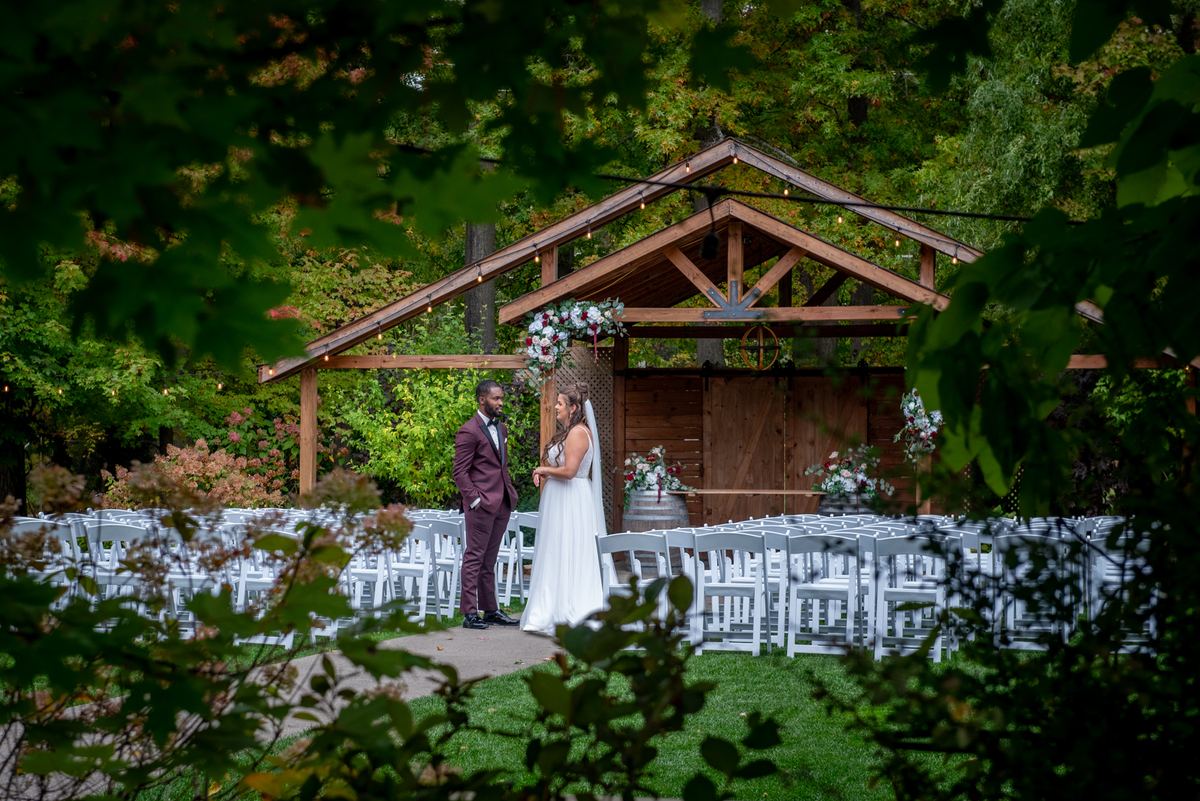 Alexandria and Derado stand facing each other at the altar beneath a rustic wooden archway, framed by lush green foliage.