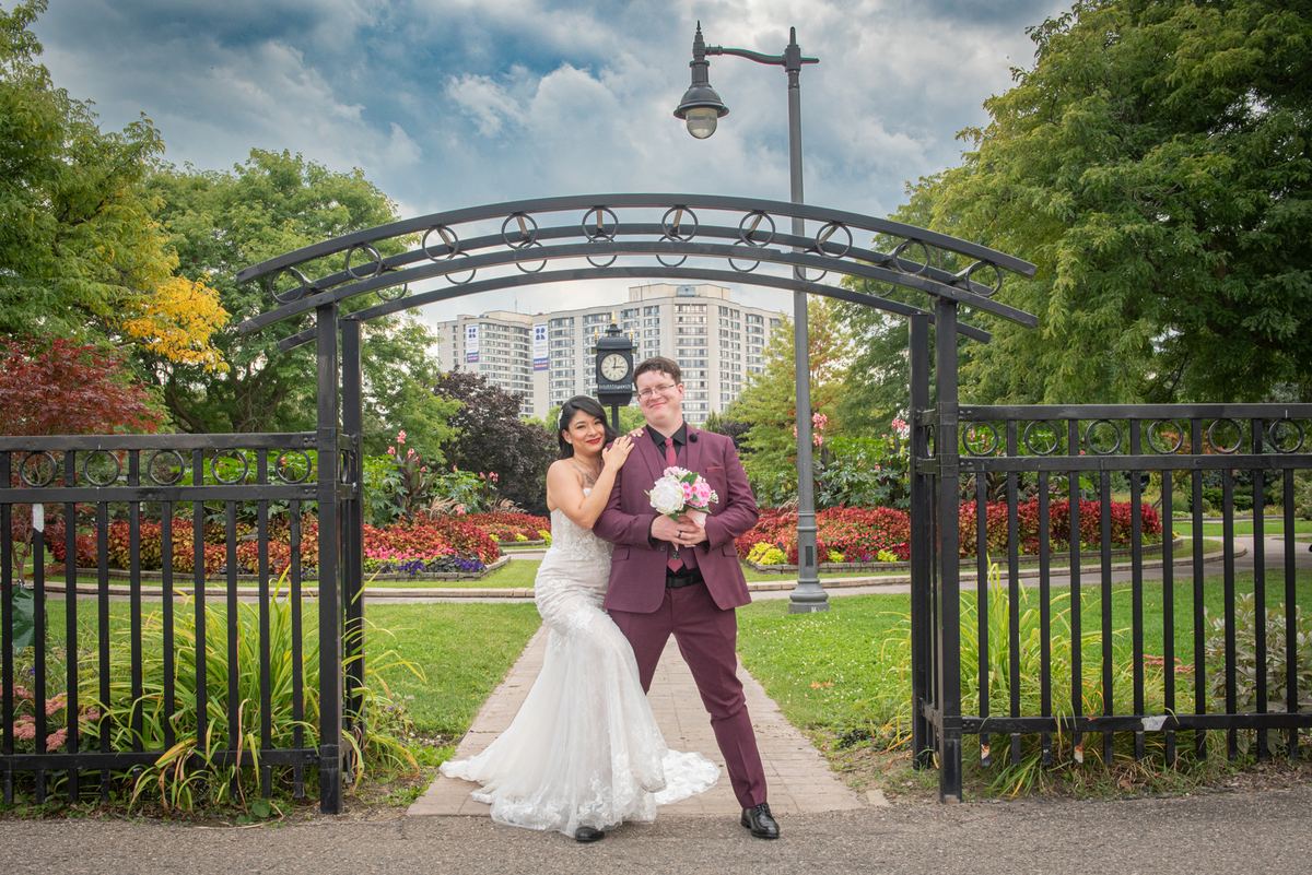 A bride in white and groom in burgundy pose under an ornate archway in a park with lush greenery and cityscape.