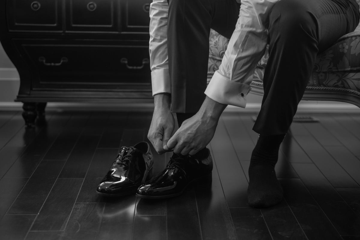 A man in formal attire ties the laces of his gleaming patent leather dress shoes on a hardwood floor.
