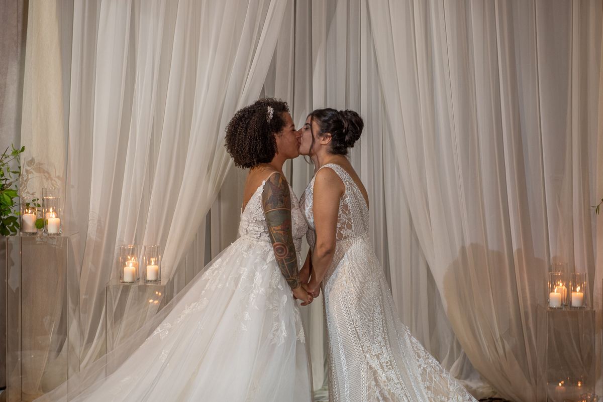 Two brides with flowing drapery behind them and candles on clear pedestals share a tender kiss while holding hands.